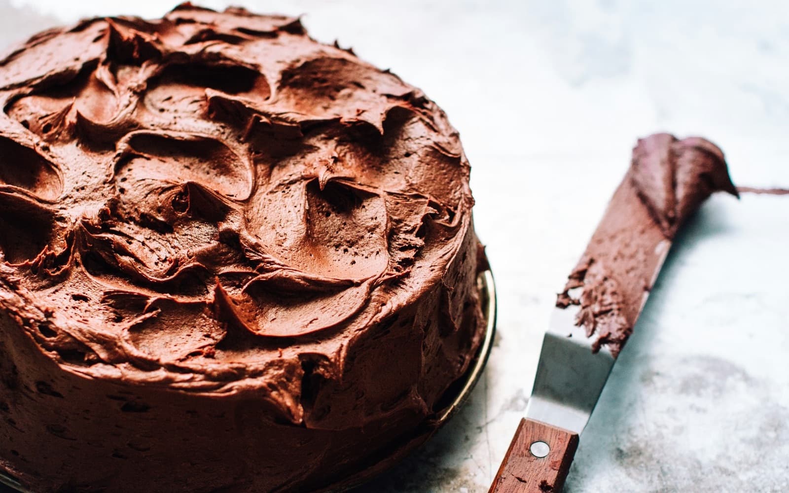 A chocolate layer cake covered in swirls of chocolate frosting, with an offset spatula beside it
